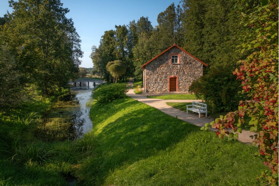 Stone house by a calm river, surrounded by green trees and grass, with a bench on a sunny day.