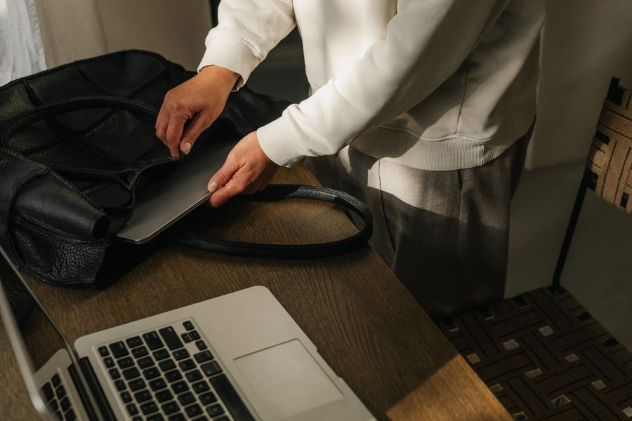 Person placing a laptop into a black bag on a wooden desk with another laptop nearby.