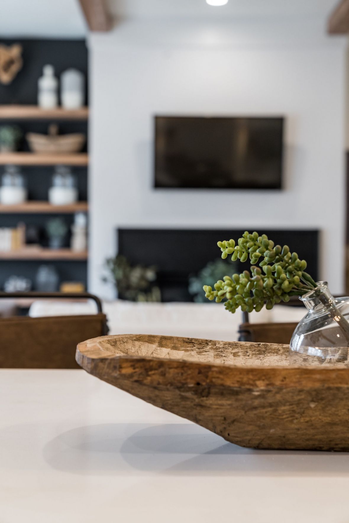 A wooden bowl with a glass vase of green succulents on a white table, blurred living room in background.